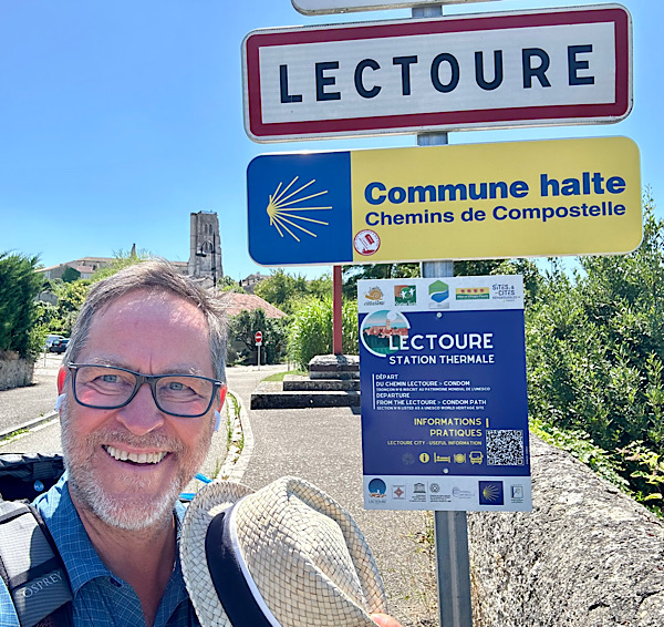 Mann mit Bart und Brille (Martin) zieht den Hut vor dem Ortsschild "Lectoure" und dem Hinweis "Commune halte, Chemins de Compostelle"