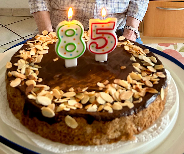 Kuchen mit Schokoladenguss und einem Kranz Mandeln aussen herum Darauf eine 85 in Form von brennenden Kerzen