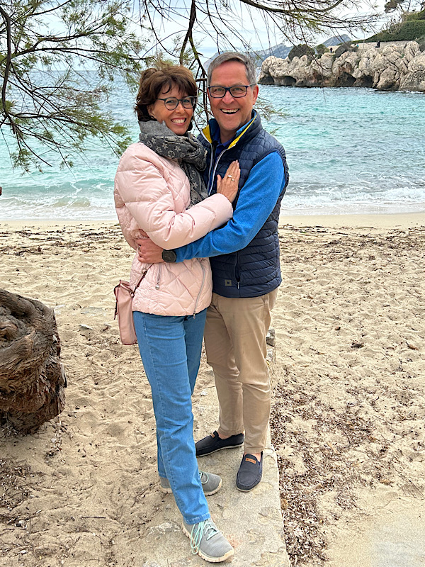 Frau mit Brille (Manu) und Mann mit Brille (Martin) stehen am Strand, umarmen sich und schauen in die Kamera