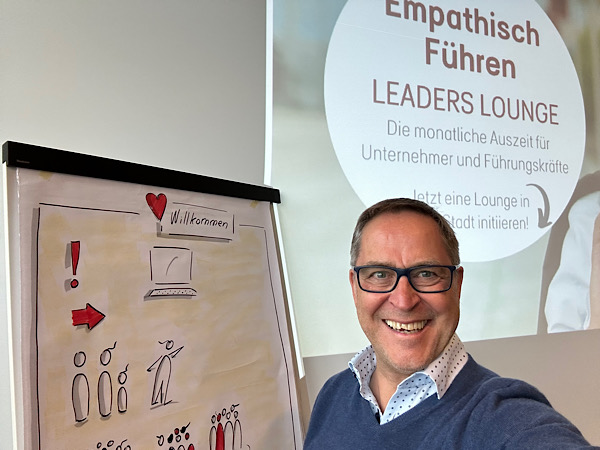 Mann (Martin) steht neben einem Flipchart und lacht in die Kamera. Im Hintergrund steht "Empathisch Führen - Leaders Lounge"