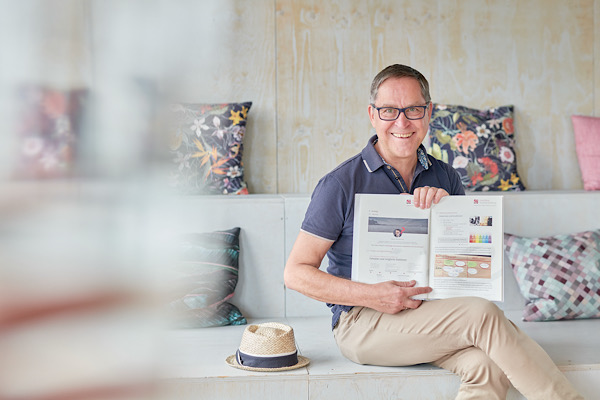 Mann mit Brille (Martin) sitzt zwischen binten Kissen auf einer Bank und zeigt ein Buch mit Text und Bildern